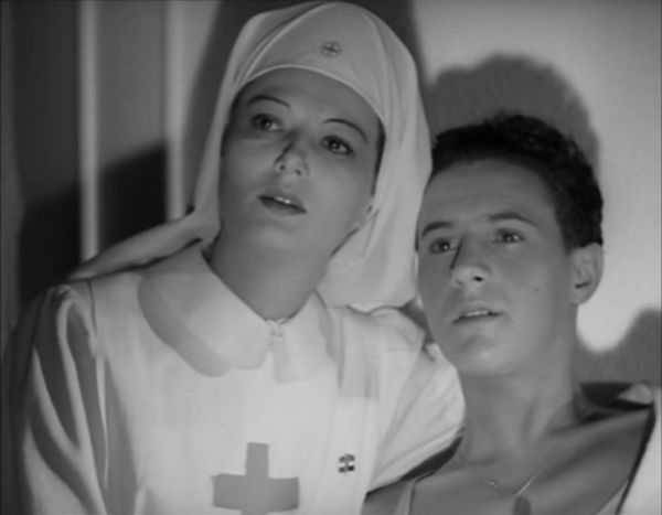 Within a room in a hospital, a young female in a nurse’s uniform wraps her arm around a recovering young man as they stare off into the distance.