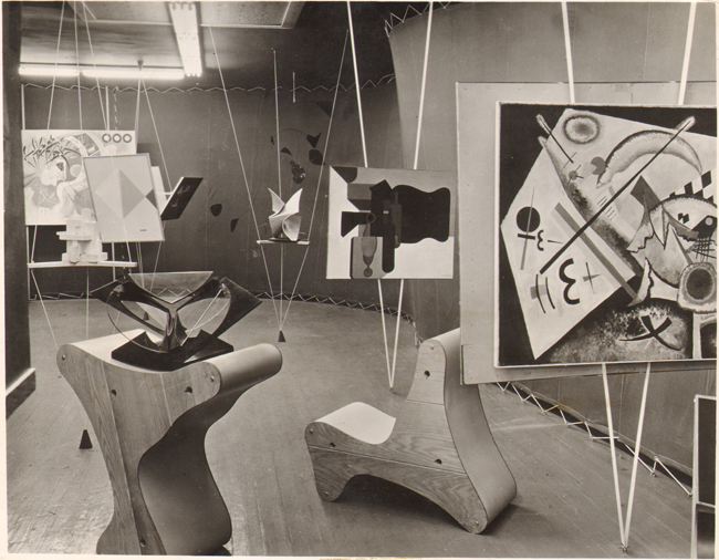 The exhibit highlights Frederick Kiesler’s Abstract Gallery, featuring numerous artworks suspended from top to bottom, anchored by strings to maintain their position. The display includes Wassily Kandinsky’s “White Cross,” prominently placed in the foreground on the right.