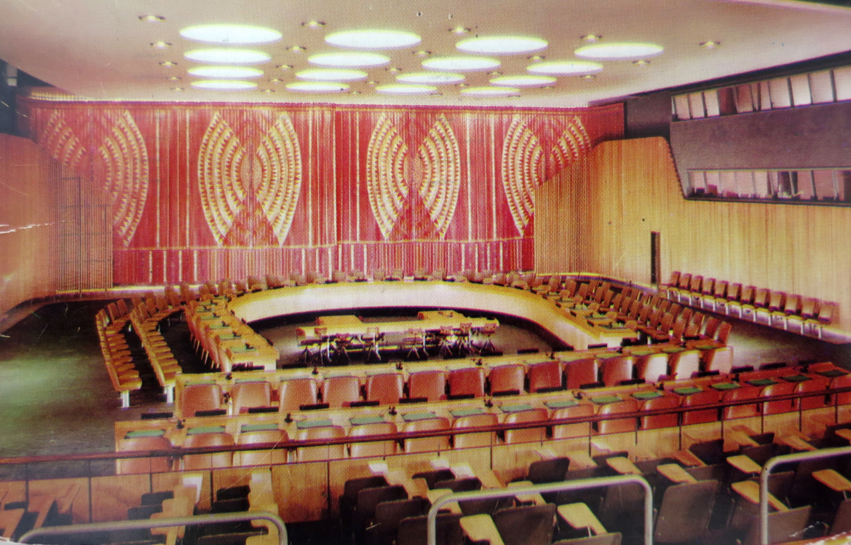 Postcard of the ECOSOC Chamber from the press gallery looking over the horseshoe table with geometric woven curtains behind it.