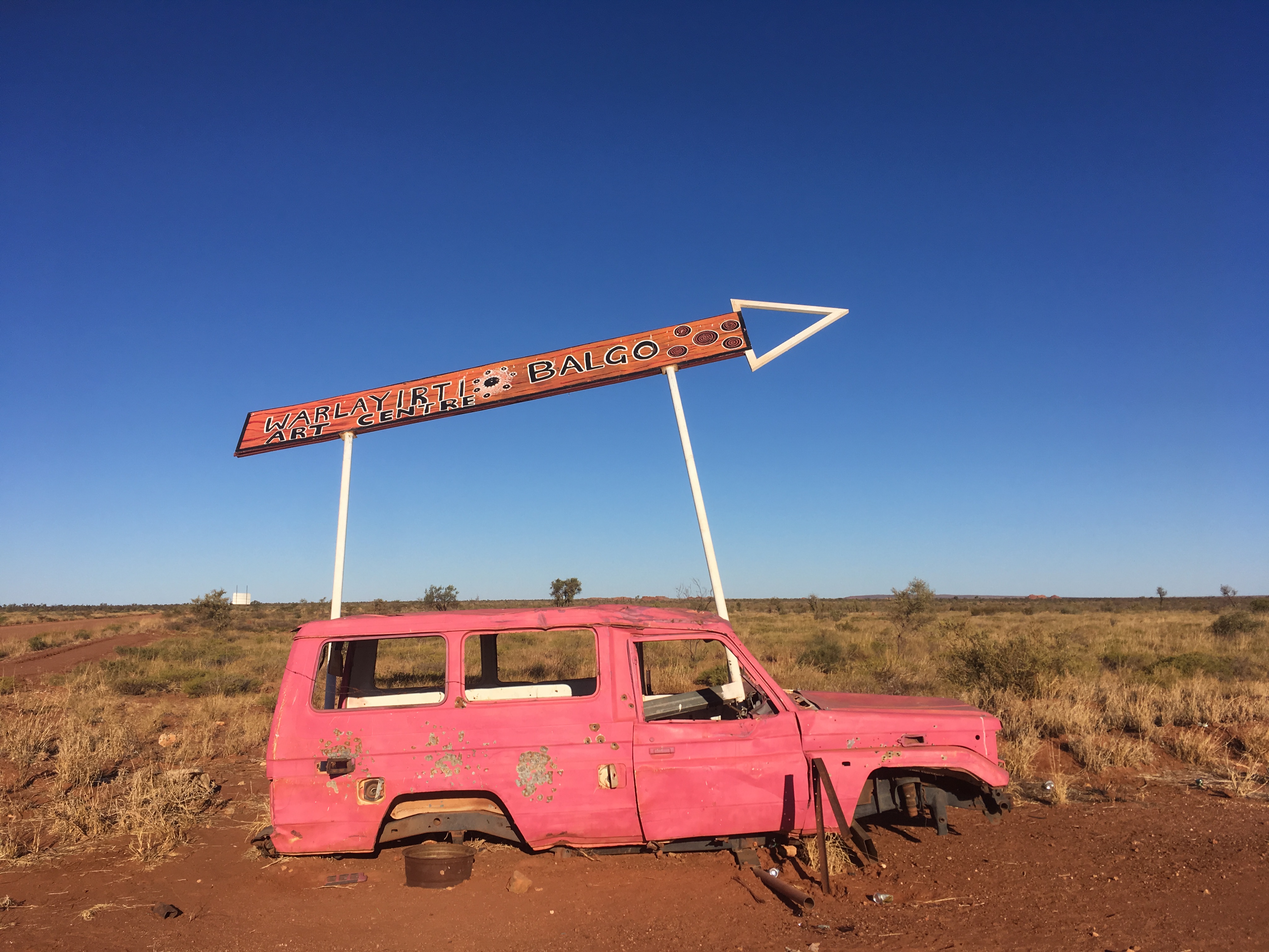 Under a clear blue sky is a pink shell of a Toyota Land Cruiser on the desert floor, with a large sign above it pointing toward Balgo.