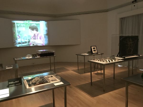 An arrangement of display tables with various materials situated atop them, each protected by glass casings. In the background a video plays against the wall.