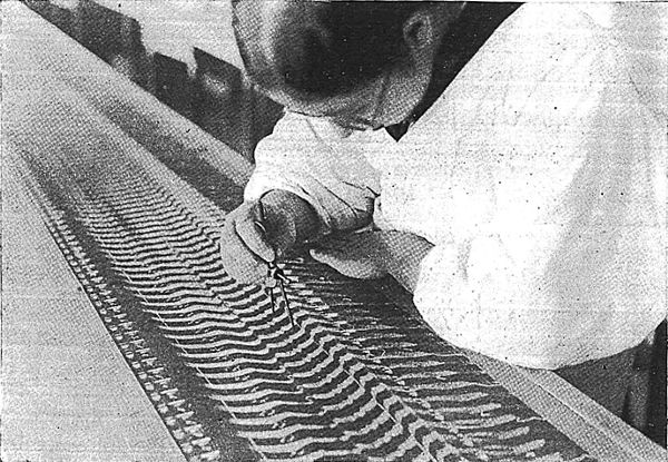 A woman in a smock with a cutting tool in her hand hunches over a composite image of several hundred of these contour lines on a long table.
