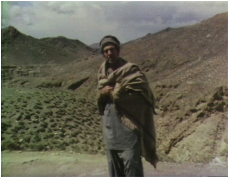 Dan Rather wearing traditional Afghani tunban and perahan looking toward the camera with a mountainous terrain behind. 