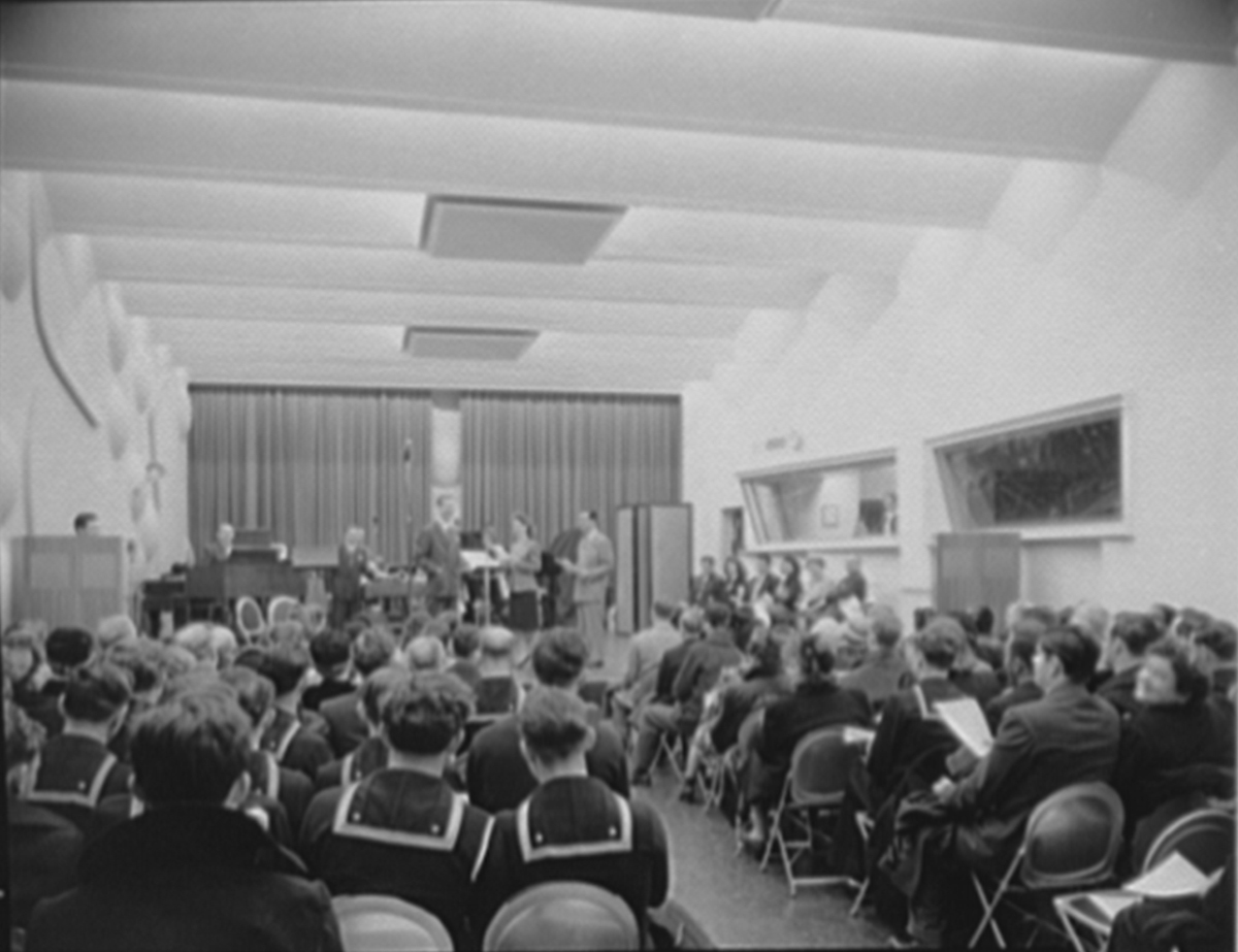 Photograph of broadcast studio interior with people on stage and a live audience of sailors, men, and women.