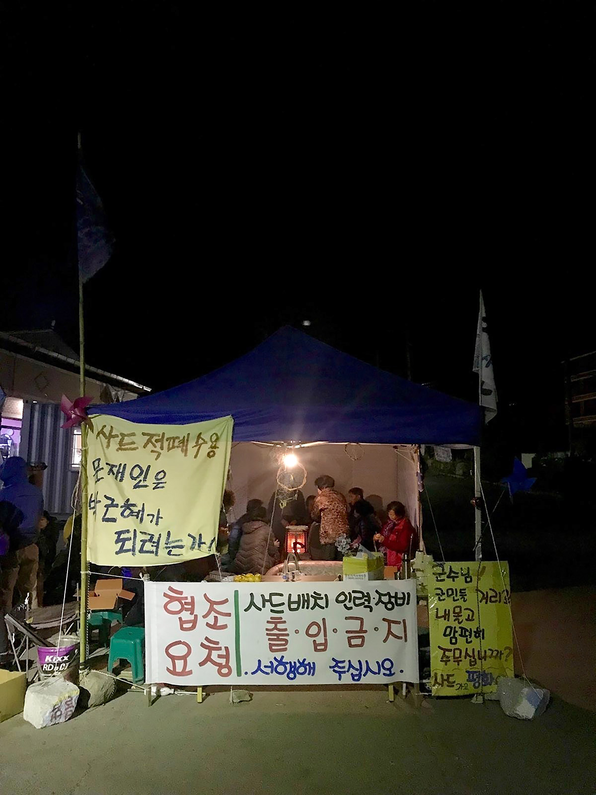 A nighttime image showing a small group of people crowded beneath a blue canopy, speaking around a portable heater. Outside the canopy is decorated with signs in Korean.
