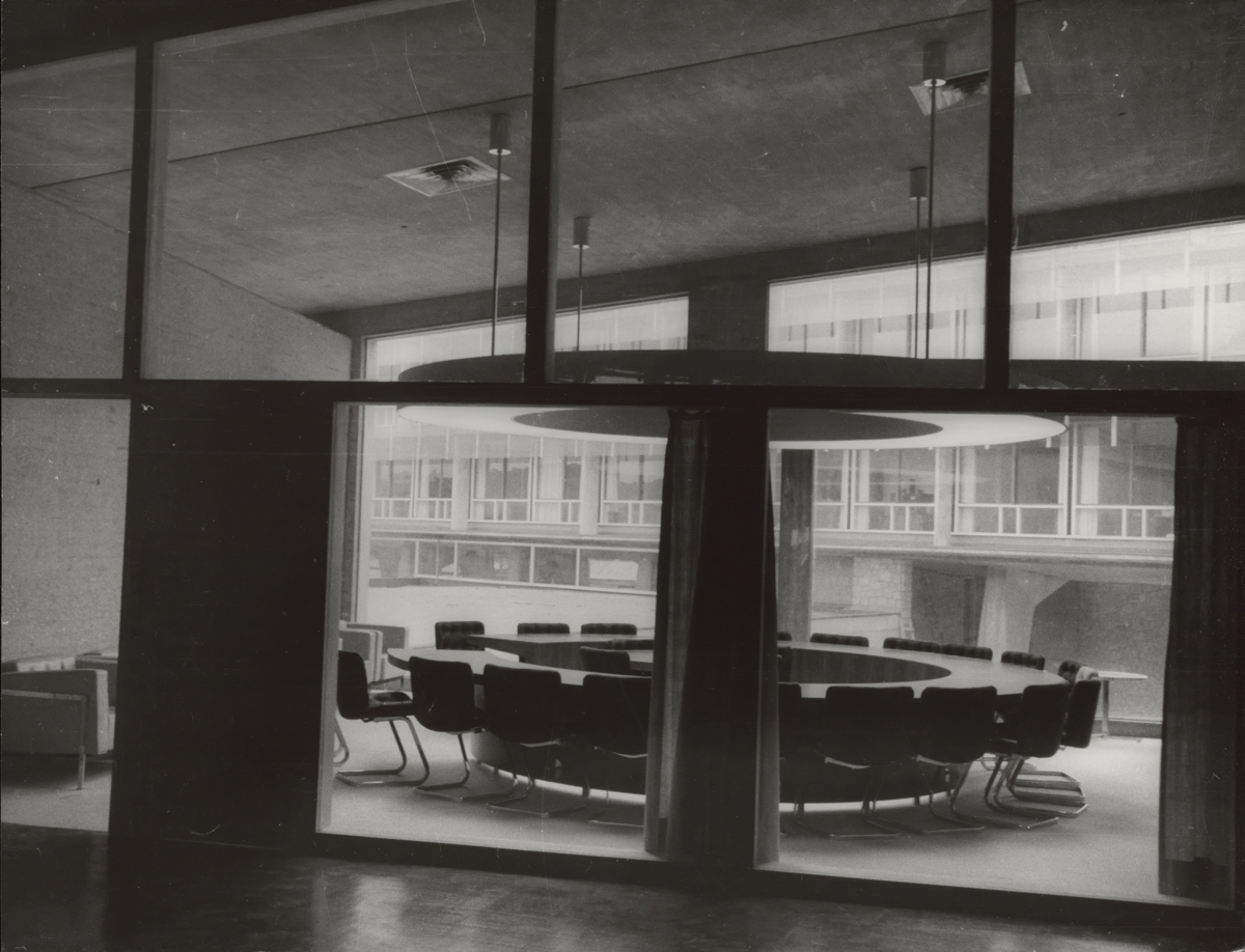 Photograph of a circular empty table with chairs and a ring of light suspended from the ceiling above it.