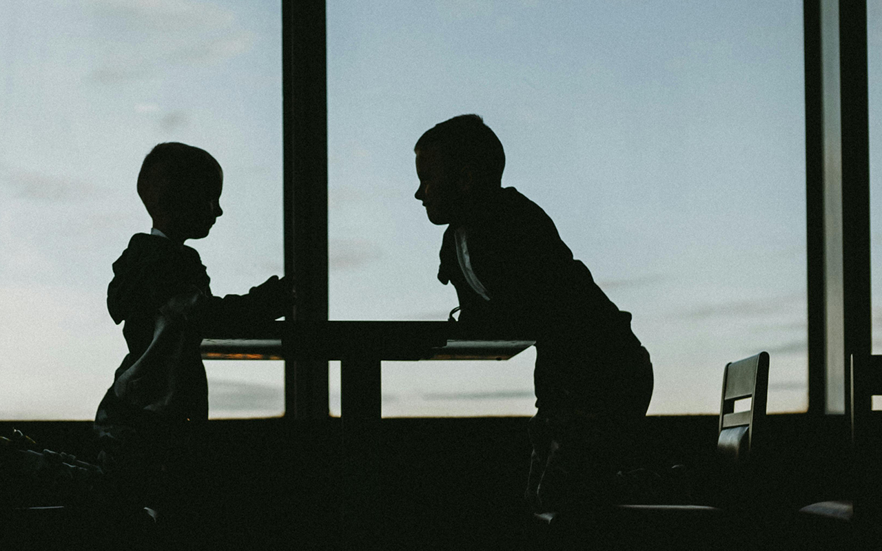 Silhouette of two children leaning on a table in front of a window opening onto a clear blue sky