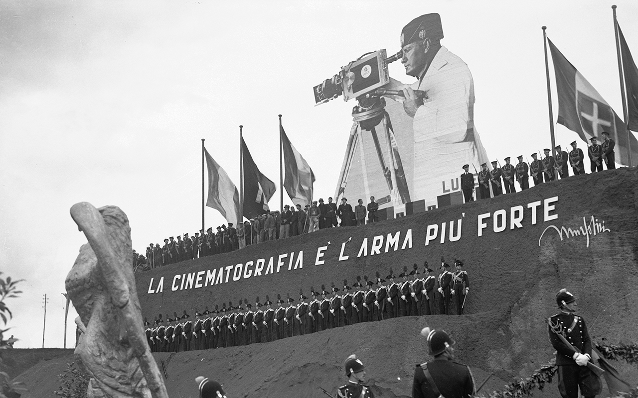 A gigantic cutout of Benito Mussolini peers into a movie camera, looming over the uniformed crowds lined up at the headquarters of the LUCE Institute.