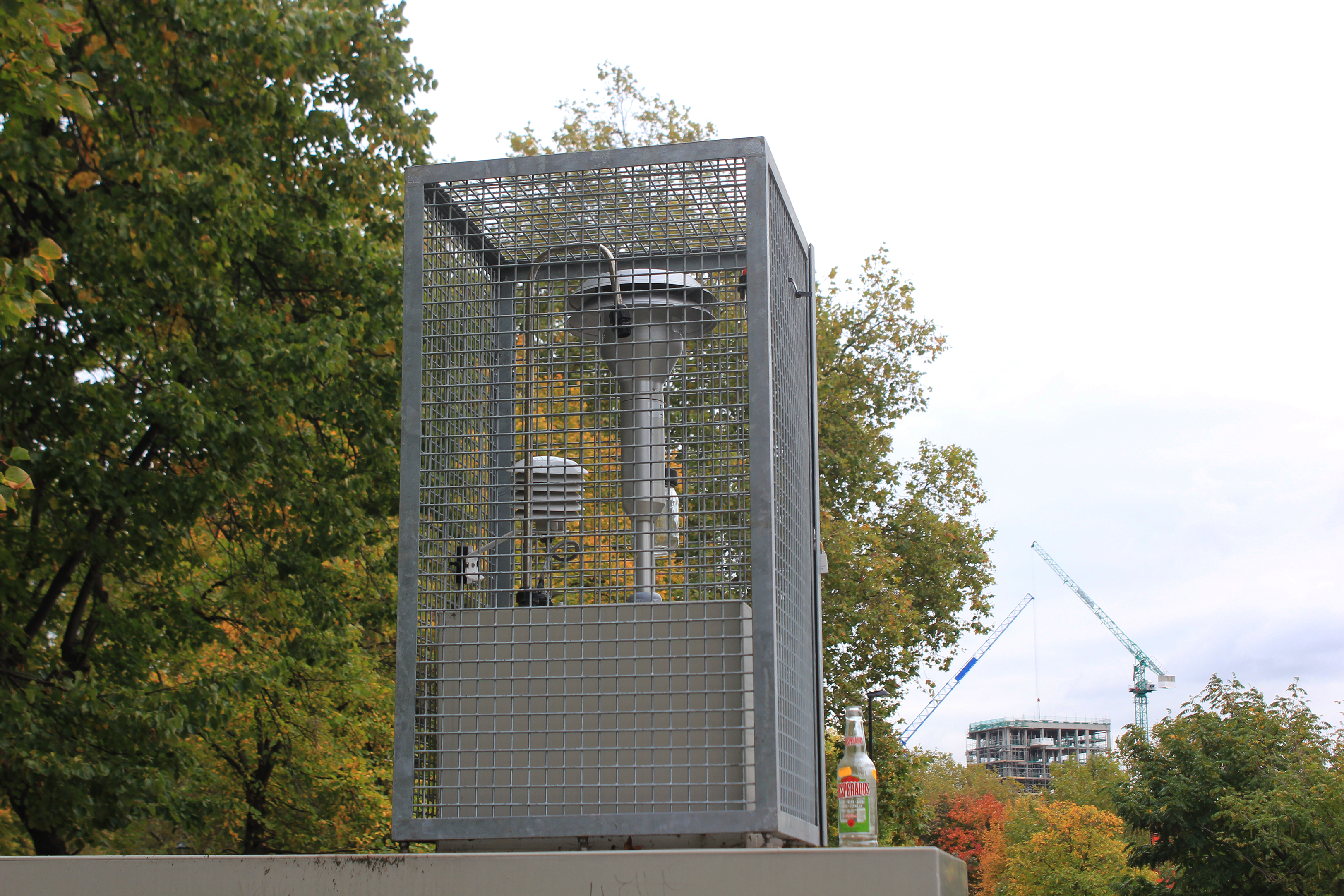 Saucer-shaped air sensor fastened atop a pole within a small fenced enclosure.