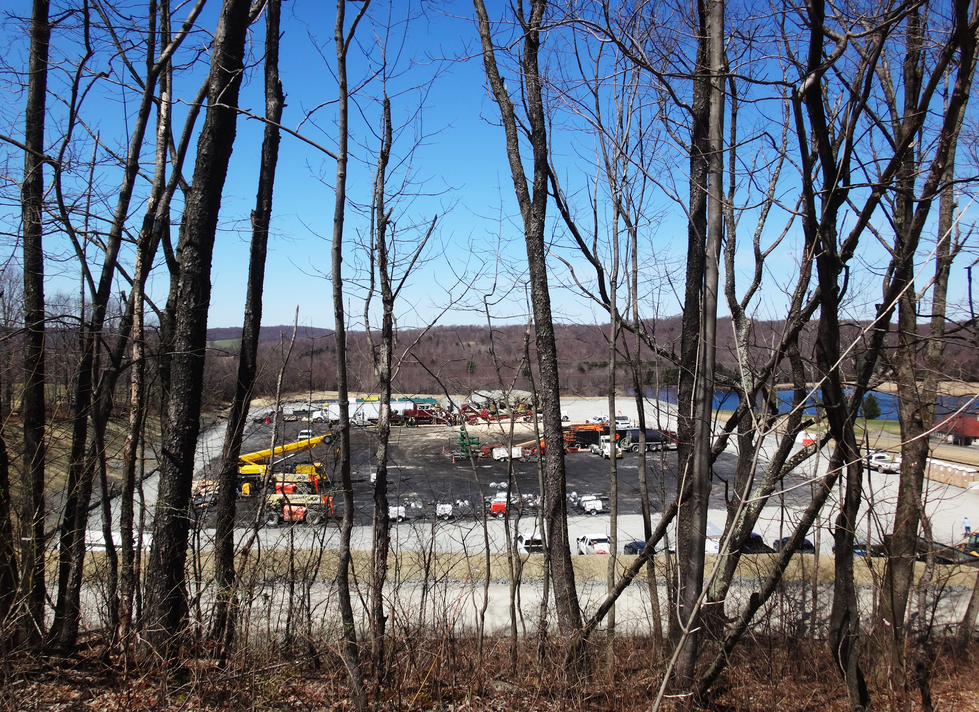 Landscape view of wooded hills in the background and a parking lot with semis, forklifts, and portable lighting stations in the foreground.