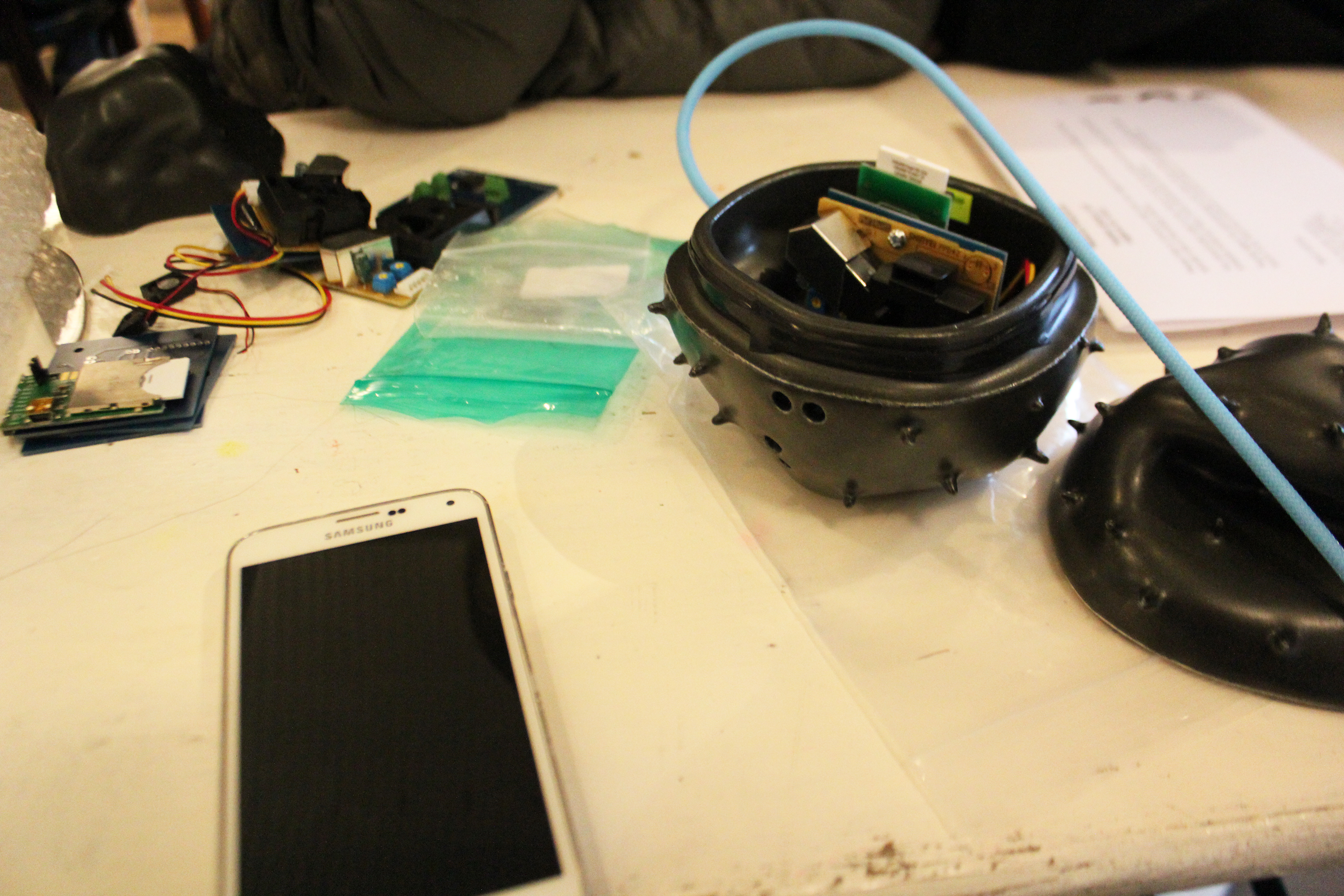 A worktable with a cellphone, loose circuit boards with wires, and a black casing with a spiny shell containing electronics.