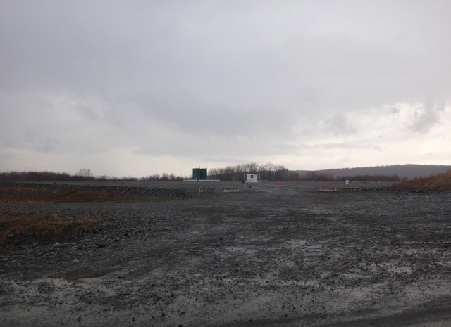 An unpaved road leading to a simple metal construction-site gate leading to two storage containers in the background.