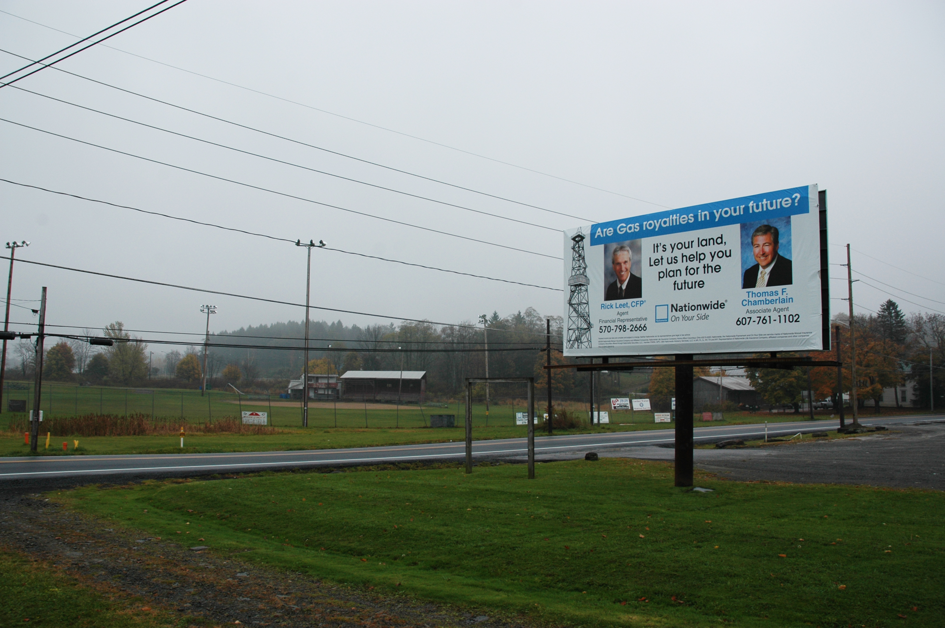 Roadside billboard beside a baseball field. The board reads, “Are Gas royalties in your future? It’s your land, Let us help you plan for the future.”