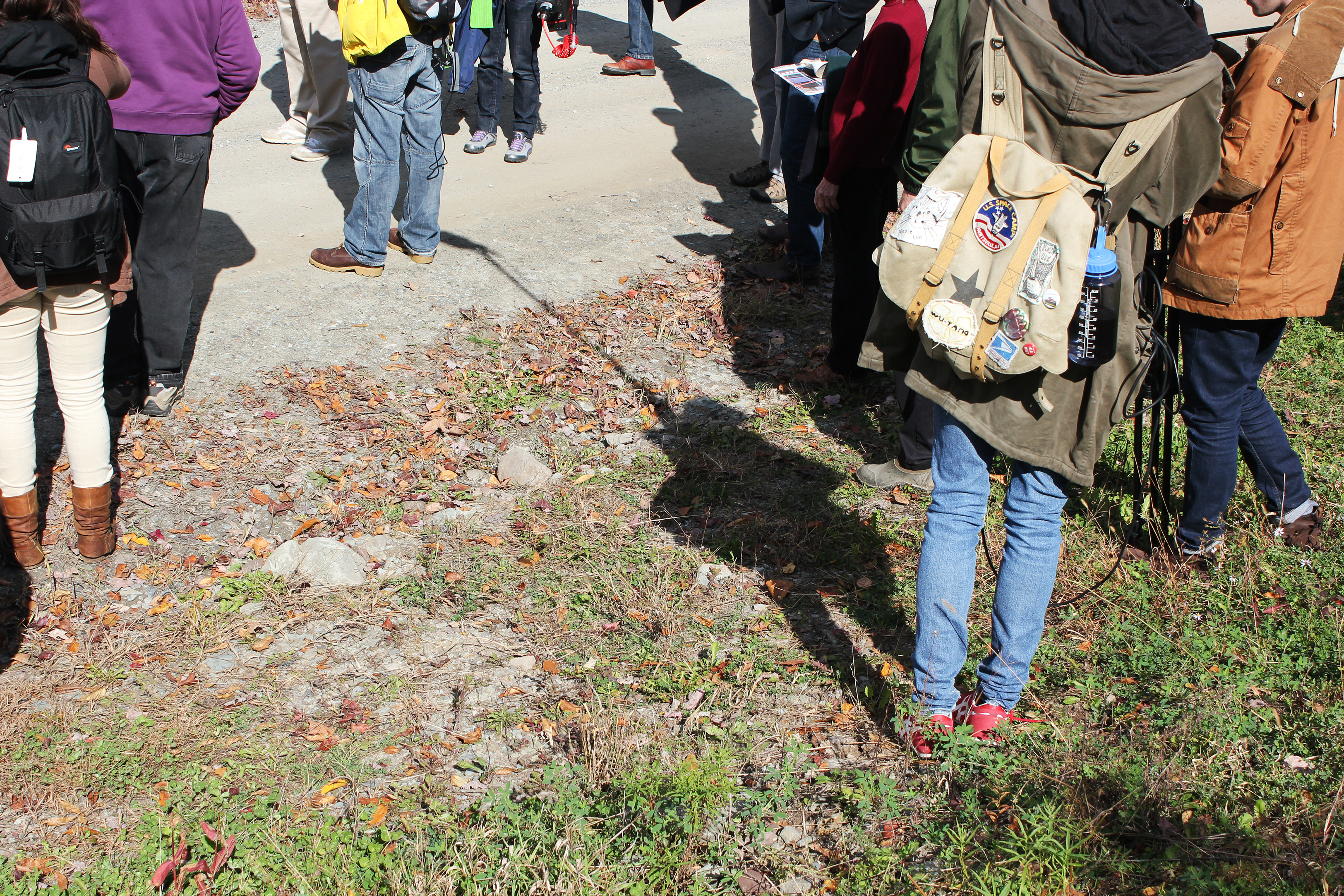 A group of people standing on or beside an unpaved road, most of whom are carrying backpacks and wearing casual wear for cool weather.