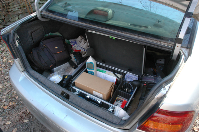 Open car trunk showing an open carrying case with a box, battery, decibel meter, N95 masks, and other assorted equipment.