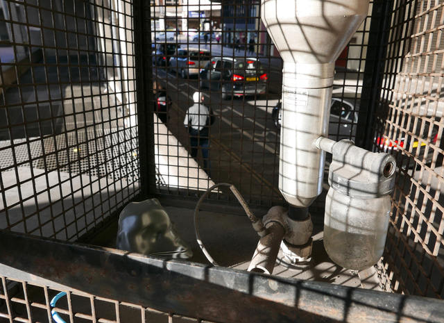 A sensor array with a black, irregular casing beside a metal air monitor, sitting within a metal cage beside a roadway.