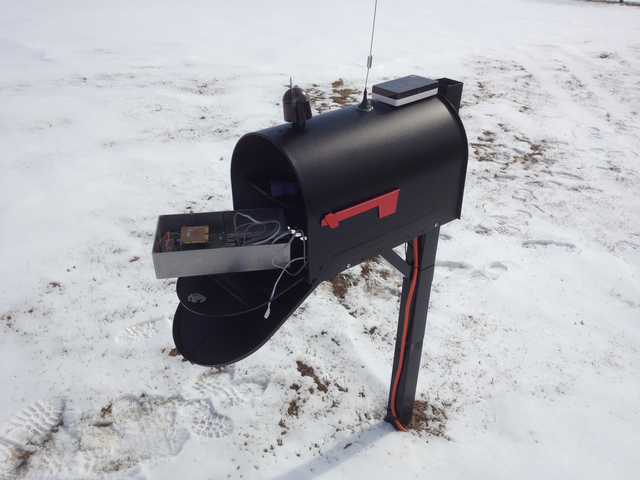 A black mailbox with a rectangular sheet metal casing with exposed wires sitting within. A wind meter and antenna are positioned on the outside.
