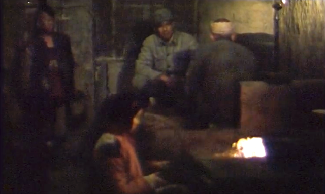 Four figures in a dark room, two are in uniform facing one another, with one wearing a head bandage. In the foreground a person tends a small file while in the background another stands against a wall.