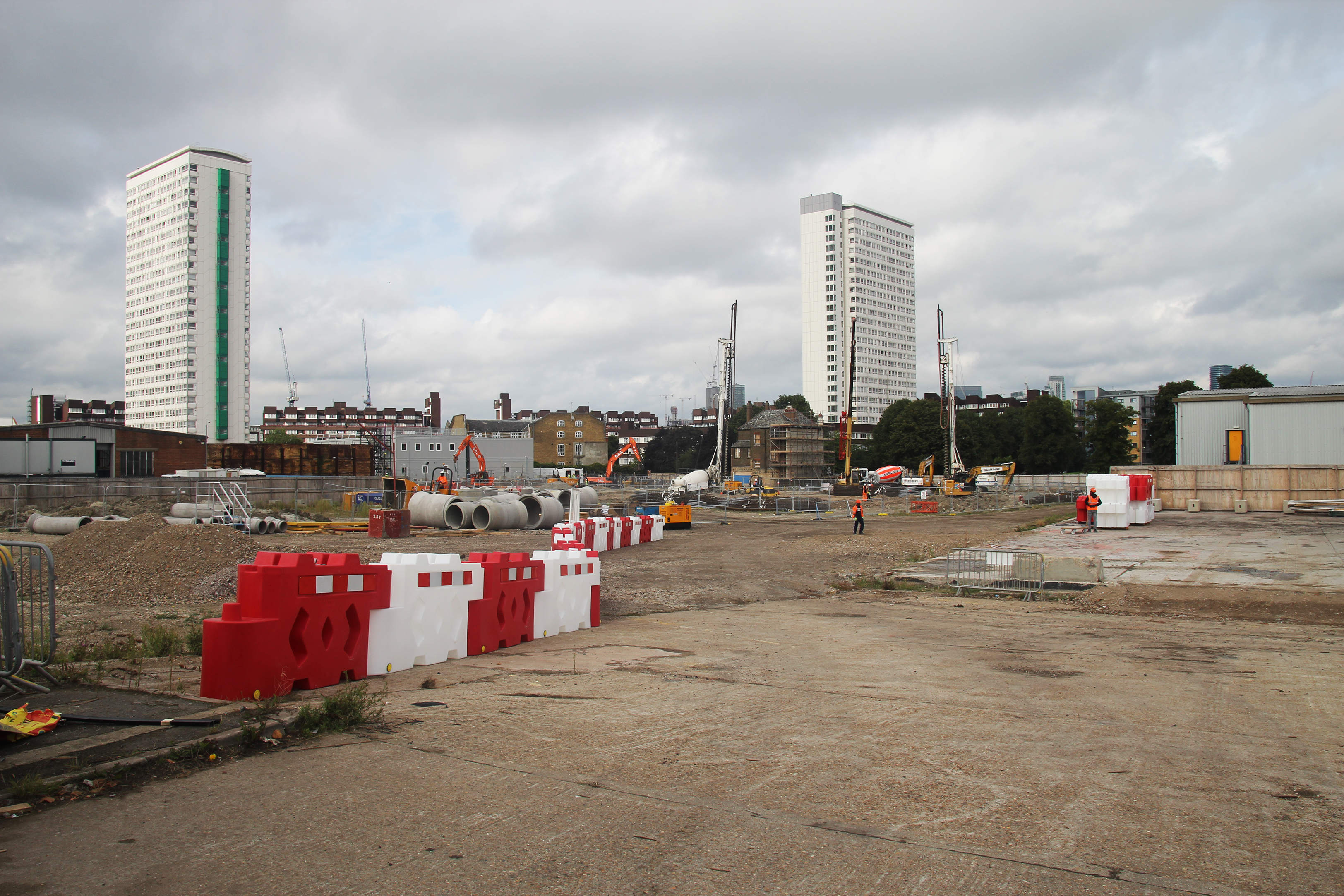 Construction site showing cleared land, concrete pipes, hazard barriers, and a handful of building cranes.