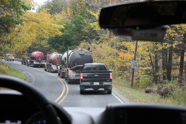 A shot of three semi trucks hauling cylindrical containers rounding a small rural road, taken from behind a dashboard.