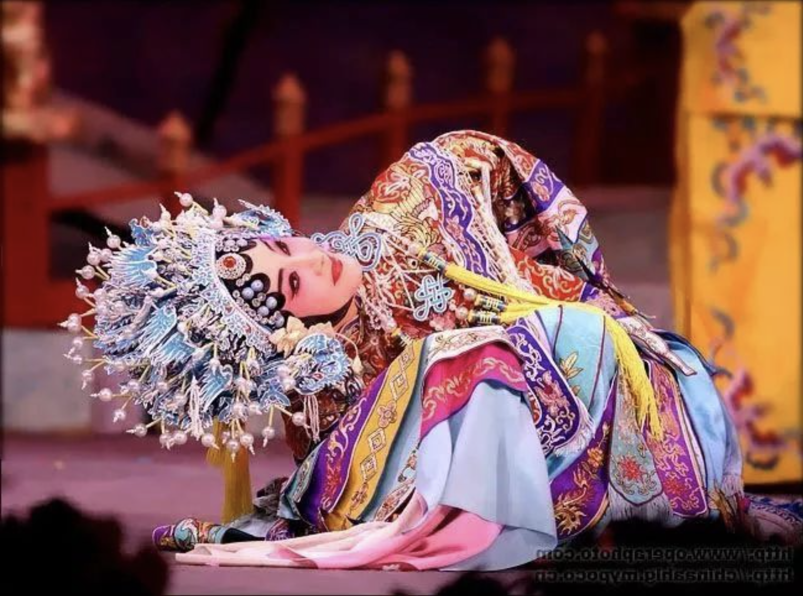 A Beijing opera actor is crouched down and leaning so far sideways that the head, neck, and torso are nearly parallel to the ground.